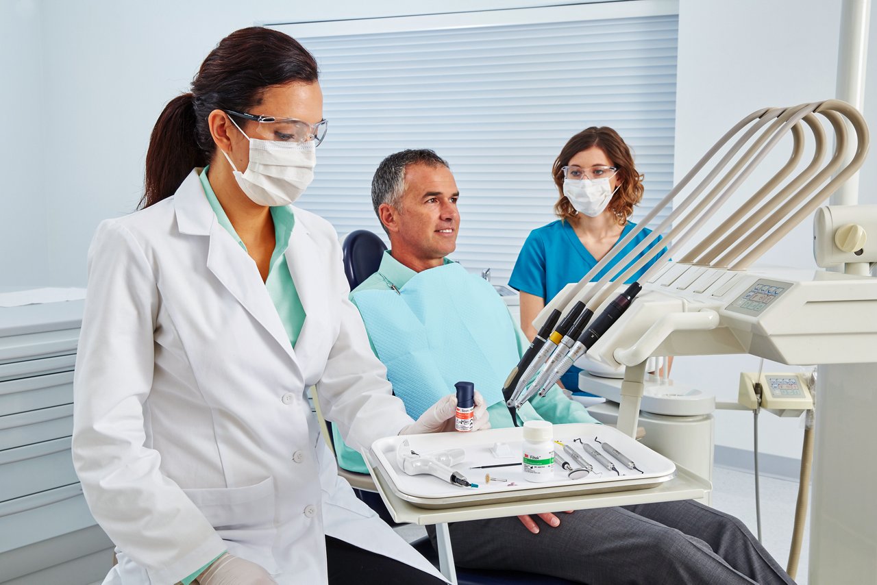 Doctor, patient and dental assistant in office room, Doctor holding a bottle of 3M™ Scotchbond™ Universal Plus Adhesive, Solventum Teal applied to shirts and SBU Plus product bottle replaced in hand. People Image, Product application, RGB, TIF