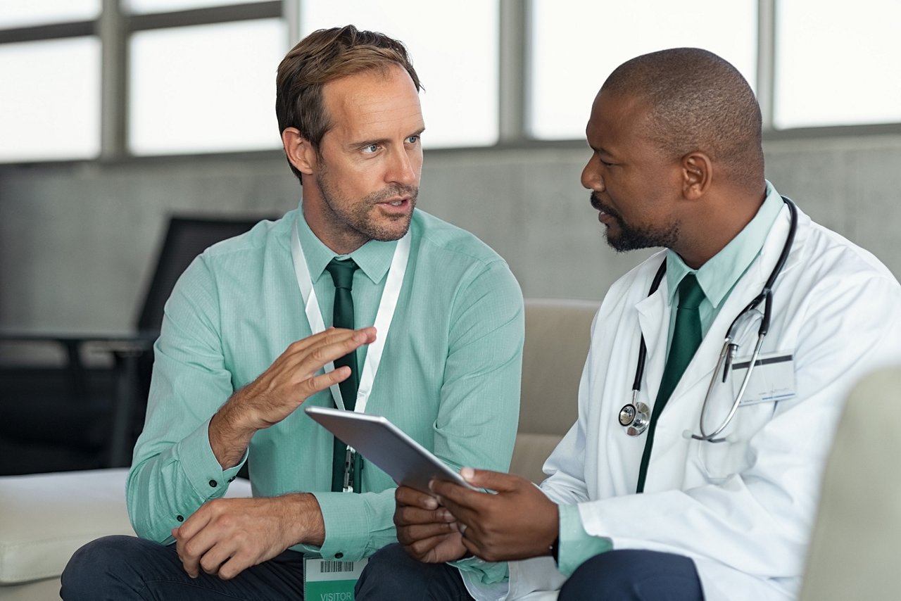 A male business leader having a seated discussion with a male clinician who is holding a tablet