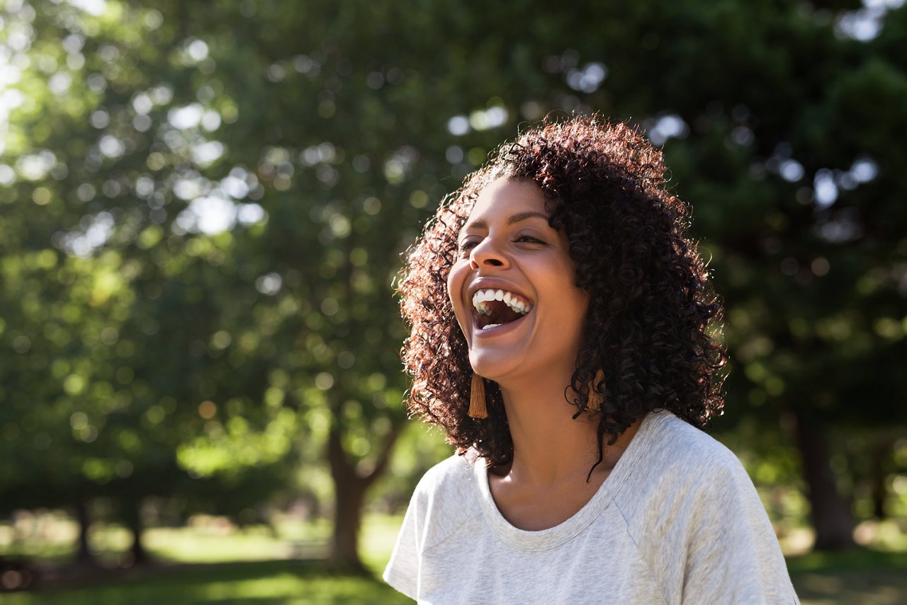 Young woman with curly hair laughing while standing outside in a park on a sunny summer afternoon; Shutwoman terstock ID 1687578475; purchase_order:credit card; job:Brand Design - Mark Bonesteel; client:mwbonesteel1@mmm.com; other:USA