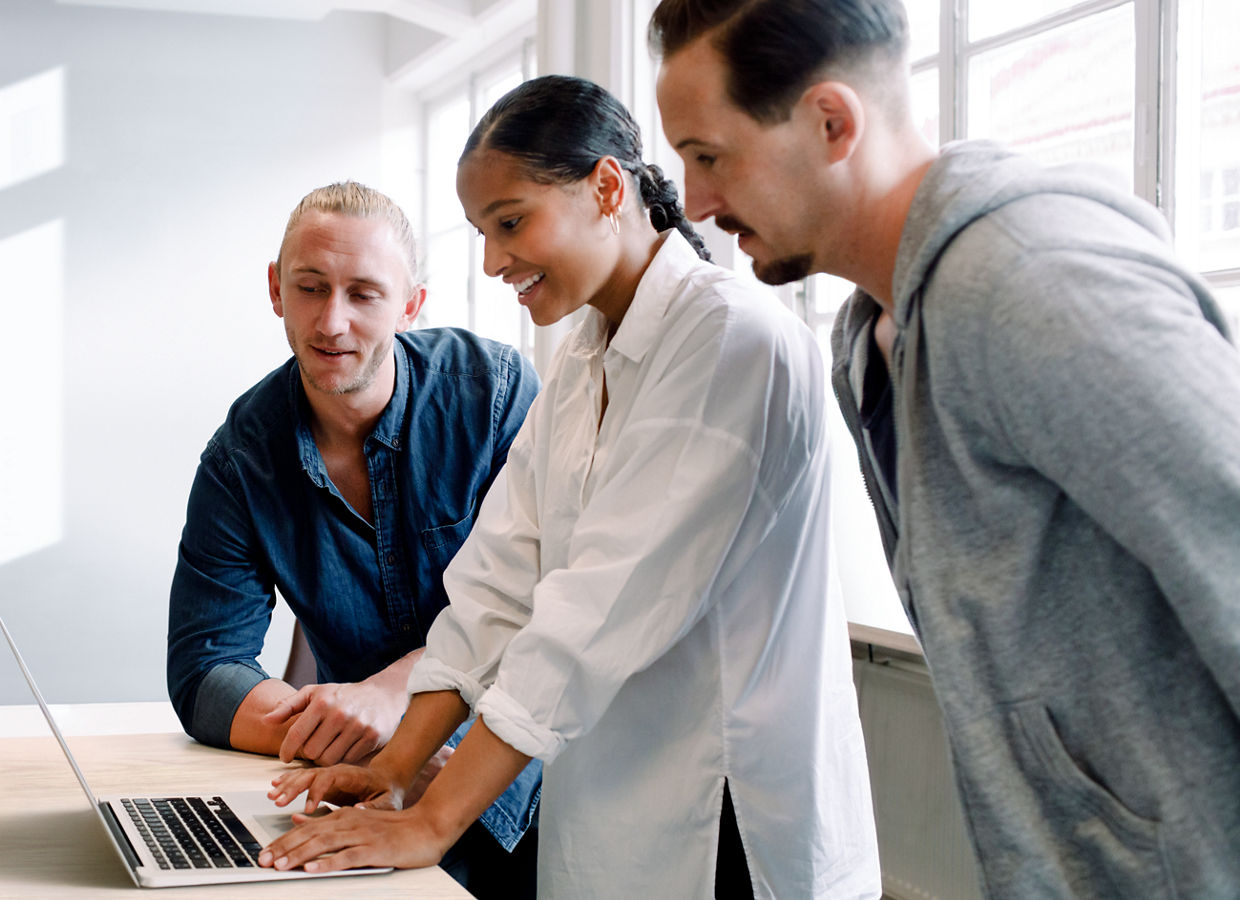 Three employees interacting with one another. A smiling female employee is working on her laptop and two men are viewing her screen.