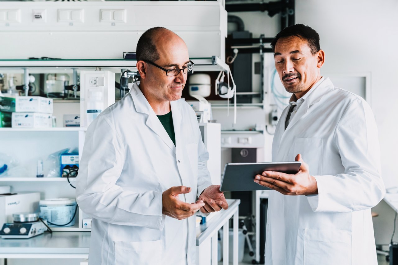 Two male scientists in lab coats interacting while viewing a tablet in a lab environment.