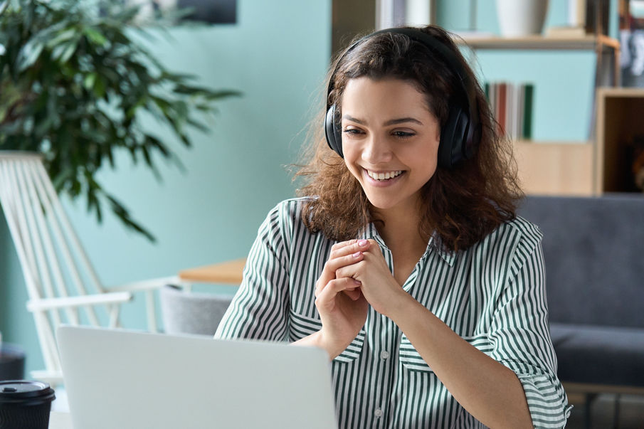 A woman wearing headphones smiling while looking at her laptop screen. 