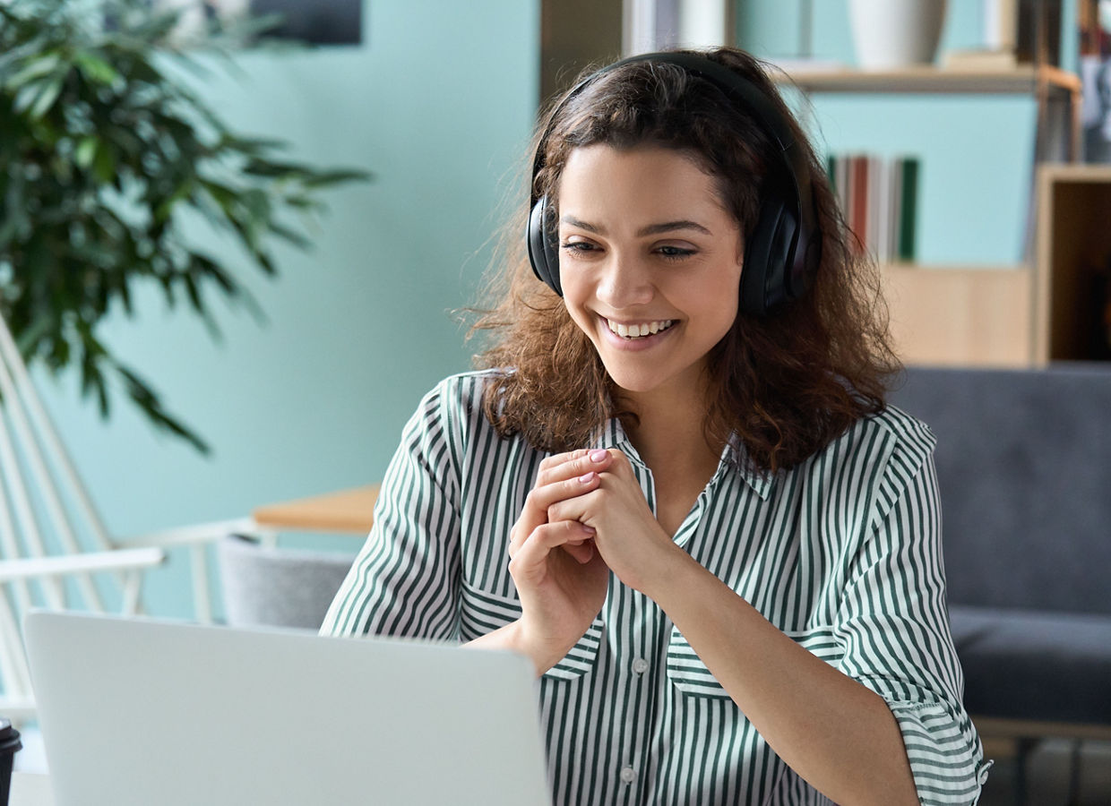 A woman wearing headphones smiling while looking at her laptop screen. 