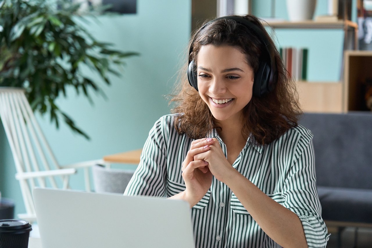 Smiling woman who is working from home. She is wearing headphones, sitting at her desk and viewing her laptop with her hands clasped in front of her.