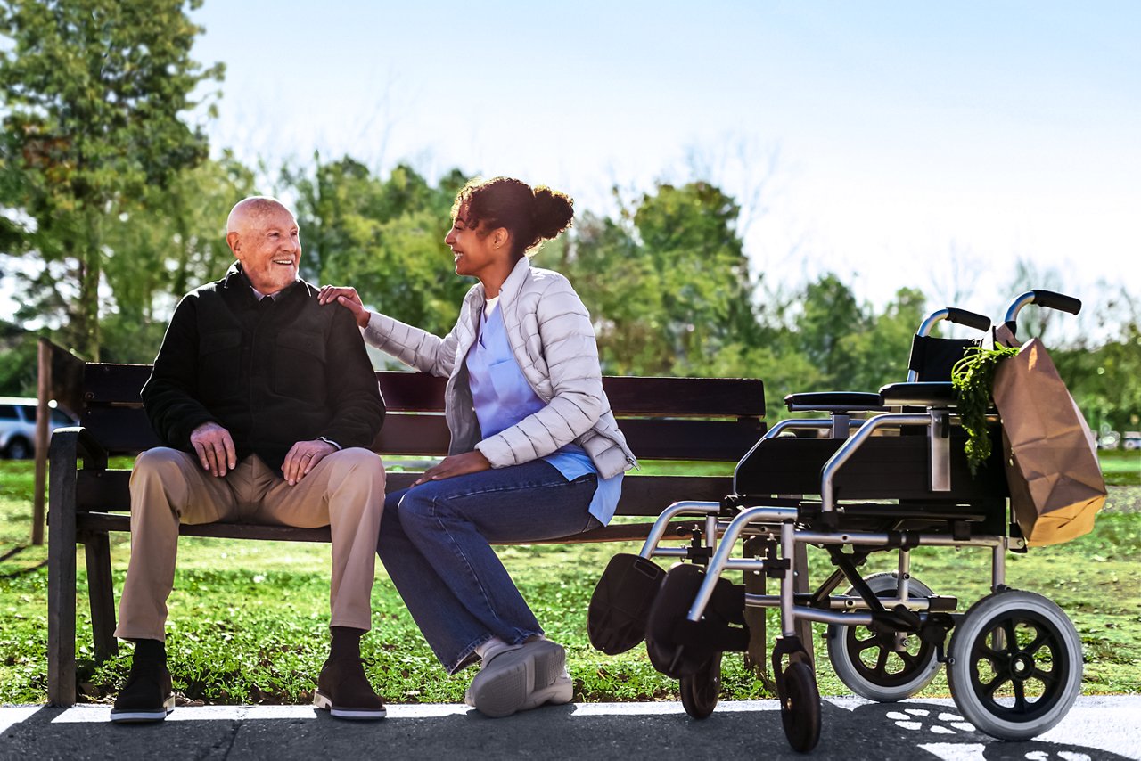 Smiling caretaker sitting on bench and talking with senior male patient in park.