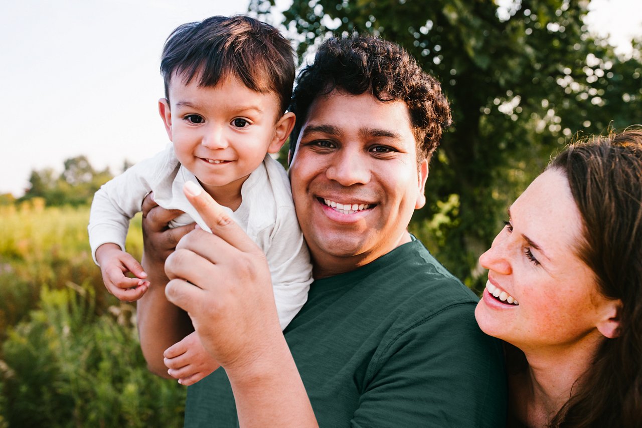 A close up of a happy couple with the man holding a baby on his shoulders in the outdoors.