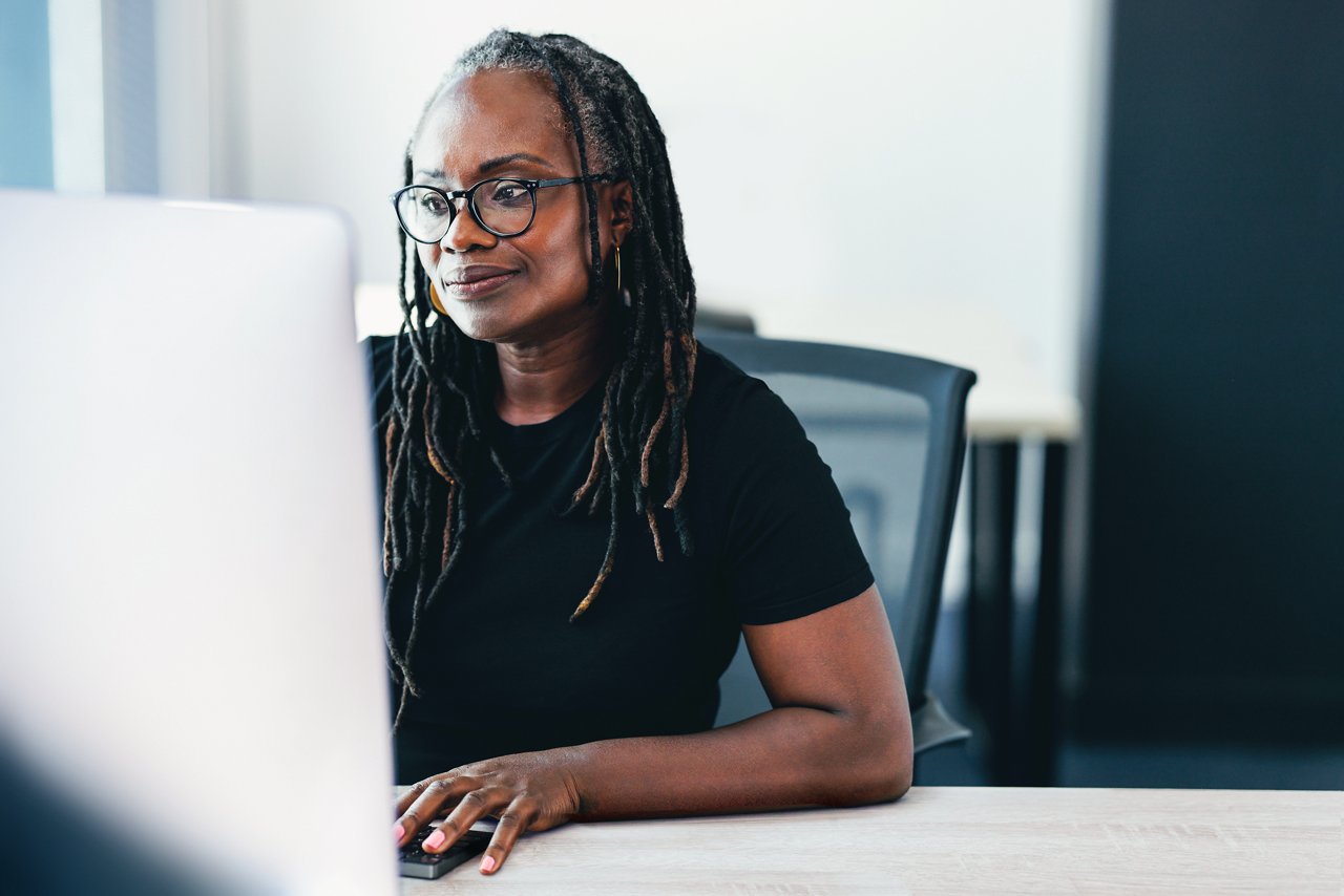 It is an image of a female office administrator seated at her desk while viewing a monitor in an office.