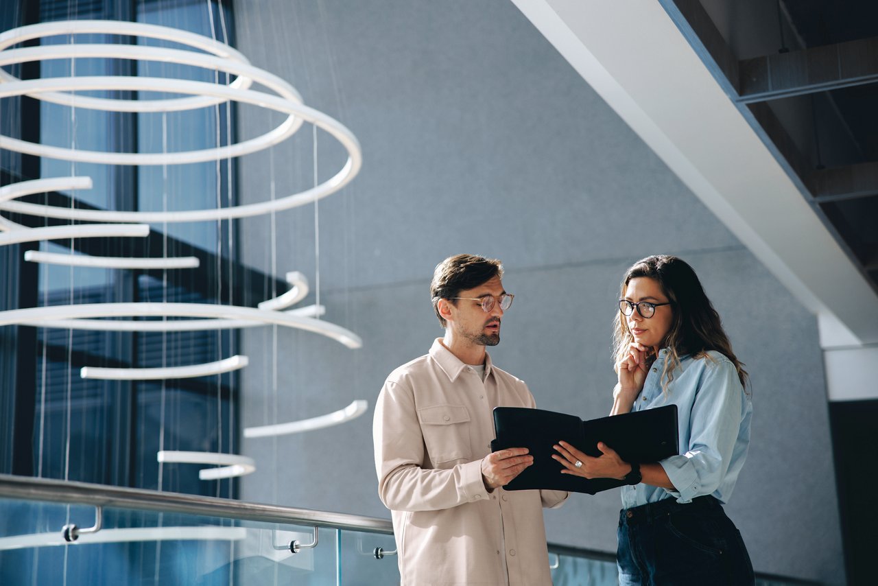 Two business people, a businessman and a businesswoman, engage in a discussion as they read a financial report together. Young business professionals working together in a modern finance company.