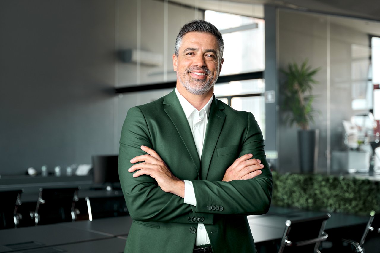 Middle-aged male employee leaning against a desk in a meeting room with his arms crossed.