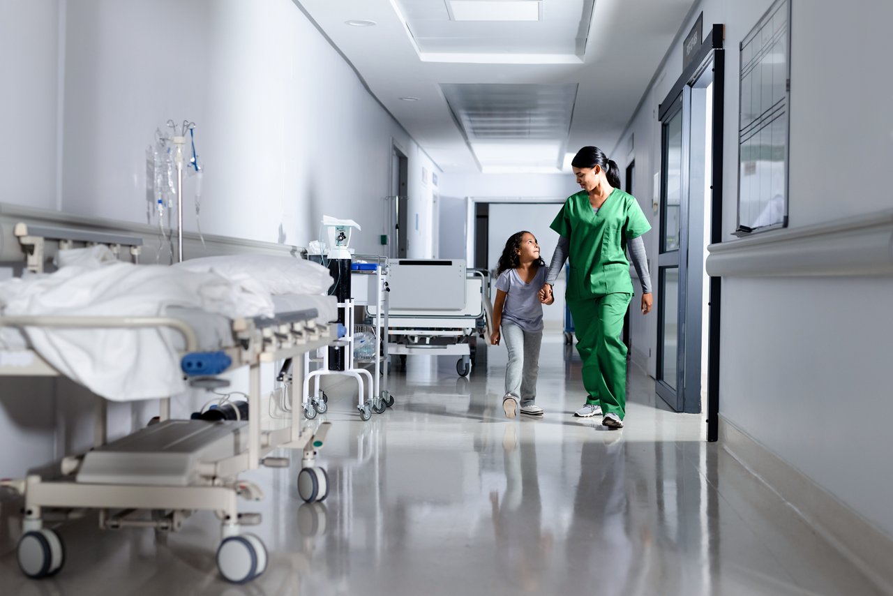 Biracial female nurse and girl patient holding hands, walking and talking in hospital corridor.