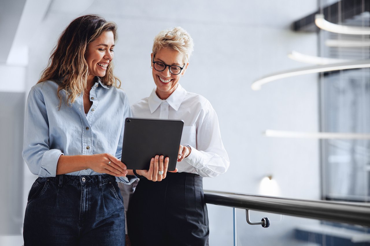 Female executive assistant holding a tablet as she shows her boss a business report indicating the company's growth and success. Happy young business woman working with a mature female professional.