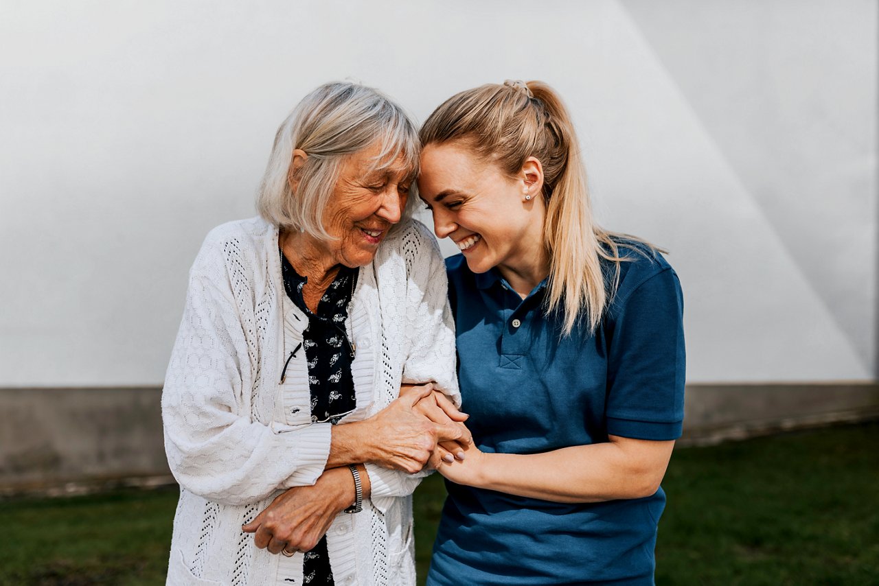 A young and older woman link arms laughing side by side, their foreheads touching.