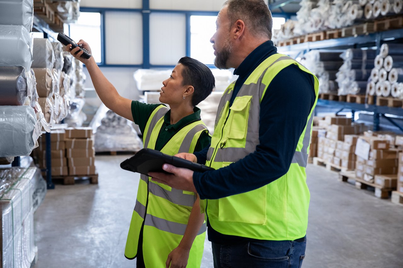 Two male workers are checking the inventory on the floor of a warehouse. One employee is holding a scanning device and the other is holding a tablet.