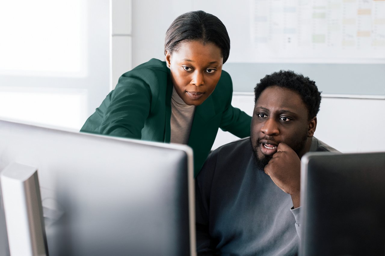 A woman standing  and pointing and a man seated, interacting while viewing a computer monitor at a desk.