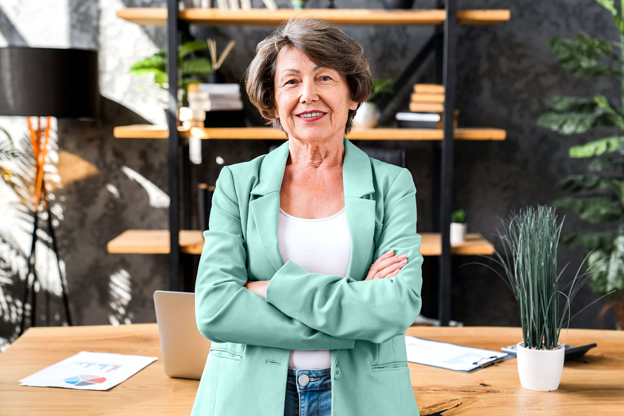 Middle-aged woman employee leaning against a desk in her office with arms crossed.