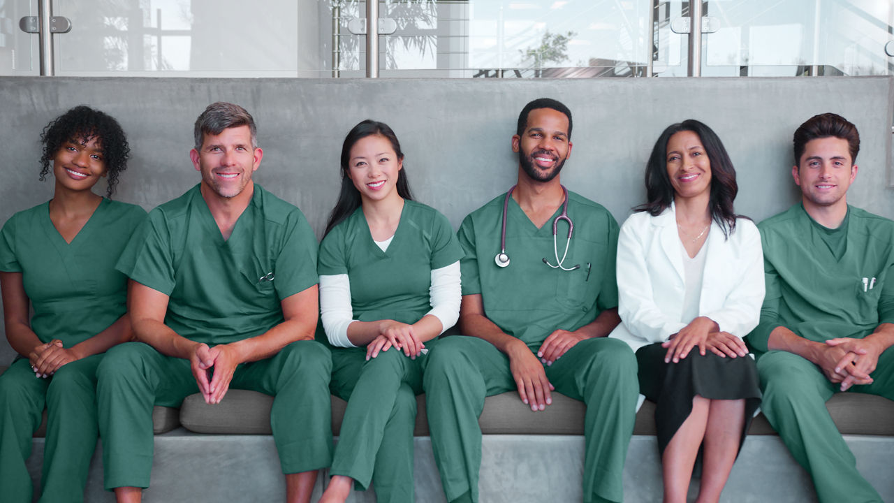 Surgical medical team in green scrubs and business attire sitting on a bench together, smiling at camera.