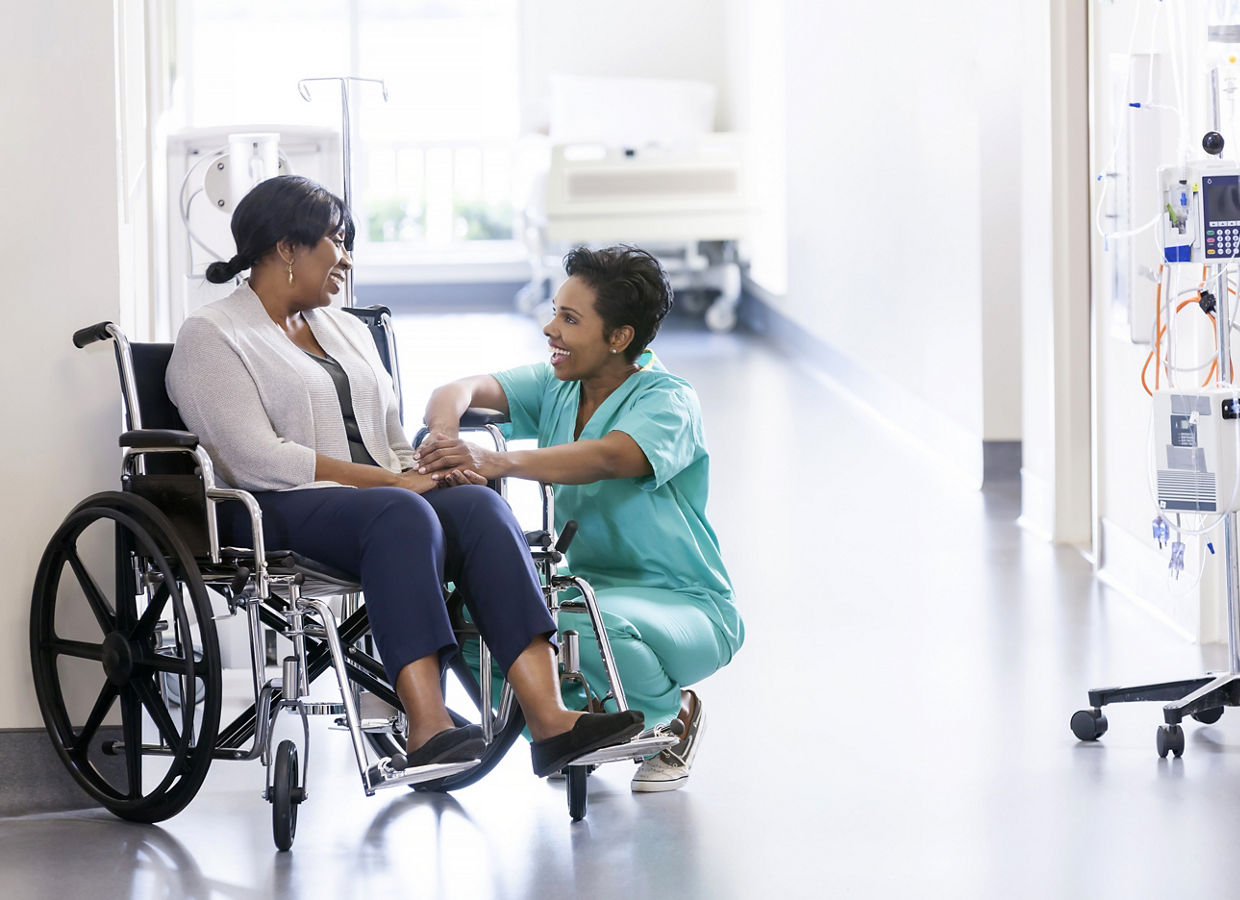 African American nurse in scrubs with patient in hospital wheelchair in corridor of specialist care clinic