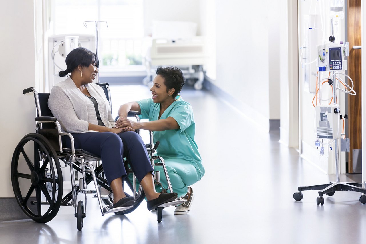 African American nurse in scrubs with patient in hospital wheelchair in corridor of specialist care clinic