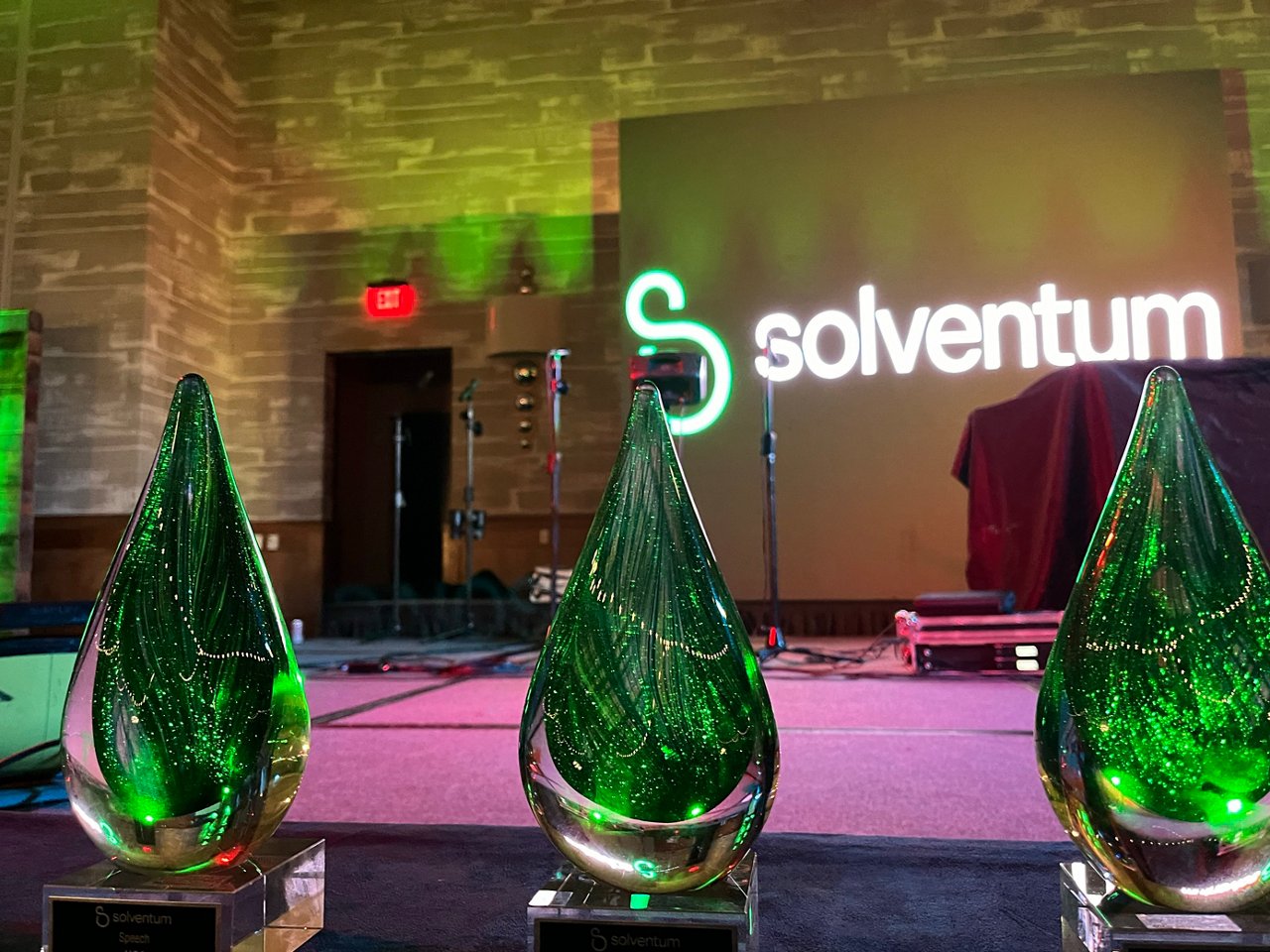 Close up of three green glass, teardrop shaped, award trophies with stage in background at the 2024 Solventum Summit Awards.