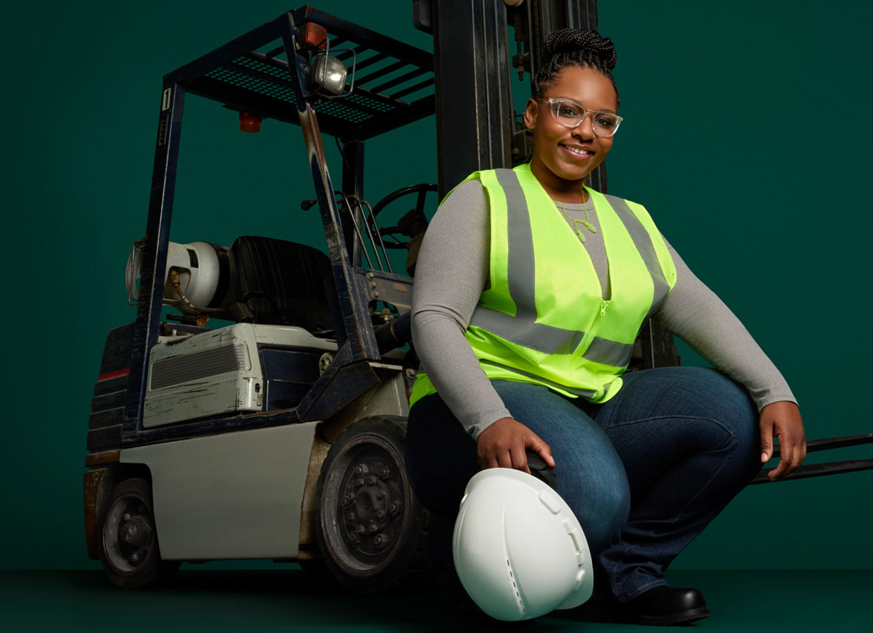 Portrait of a female manufacturing employee with hard hat and safety vest, squatting in front of forklift on a dark green background.
