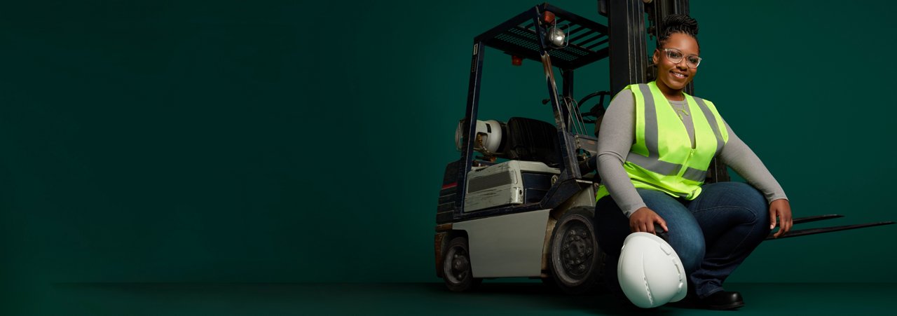 Portrait of a female manufacturing employee with hard hat and safety vest, squatting in front of forklift on a dark green background.