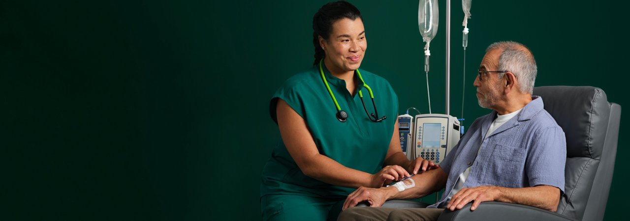 Male patient sitting in a chair with an IV attached to his arm. A smiling nurse kneels beside him checking on the patient's IV.