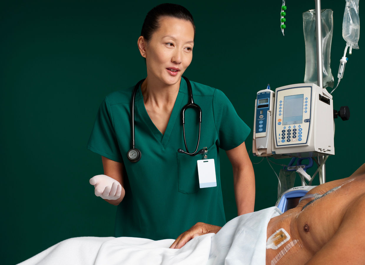 Female nurse checking on male patient in hospital bed with IVs and negative pressure wound therapy dressings applied to chest, on dark green background.