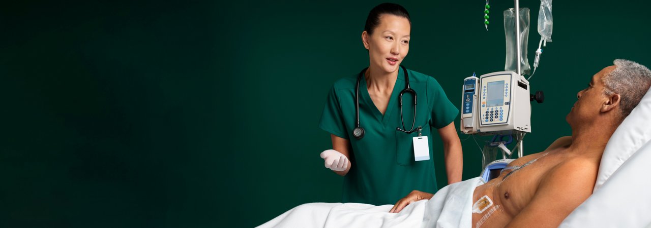 Female nurse checking on male patient in hospital bed with IVs and negative pressure wound therapy dressings applied to chest, on dark green background.