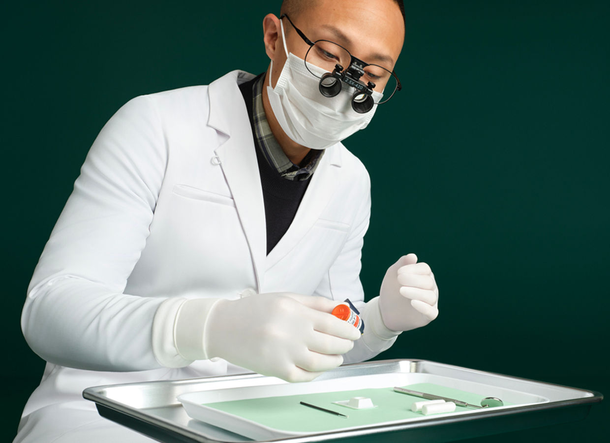 Male dentist sitting behind dental tray on stand using Scotchbond Universal Plus Adhesive, dark green background.