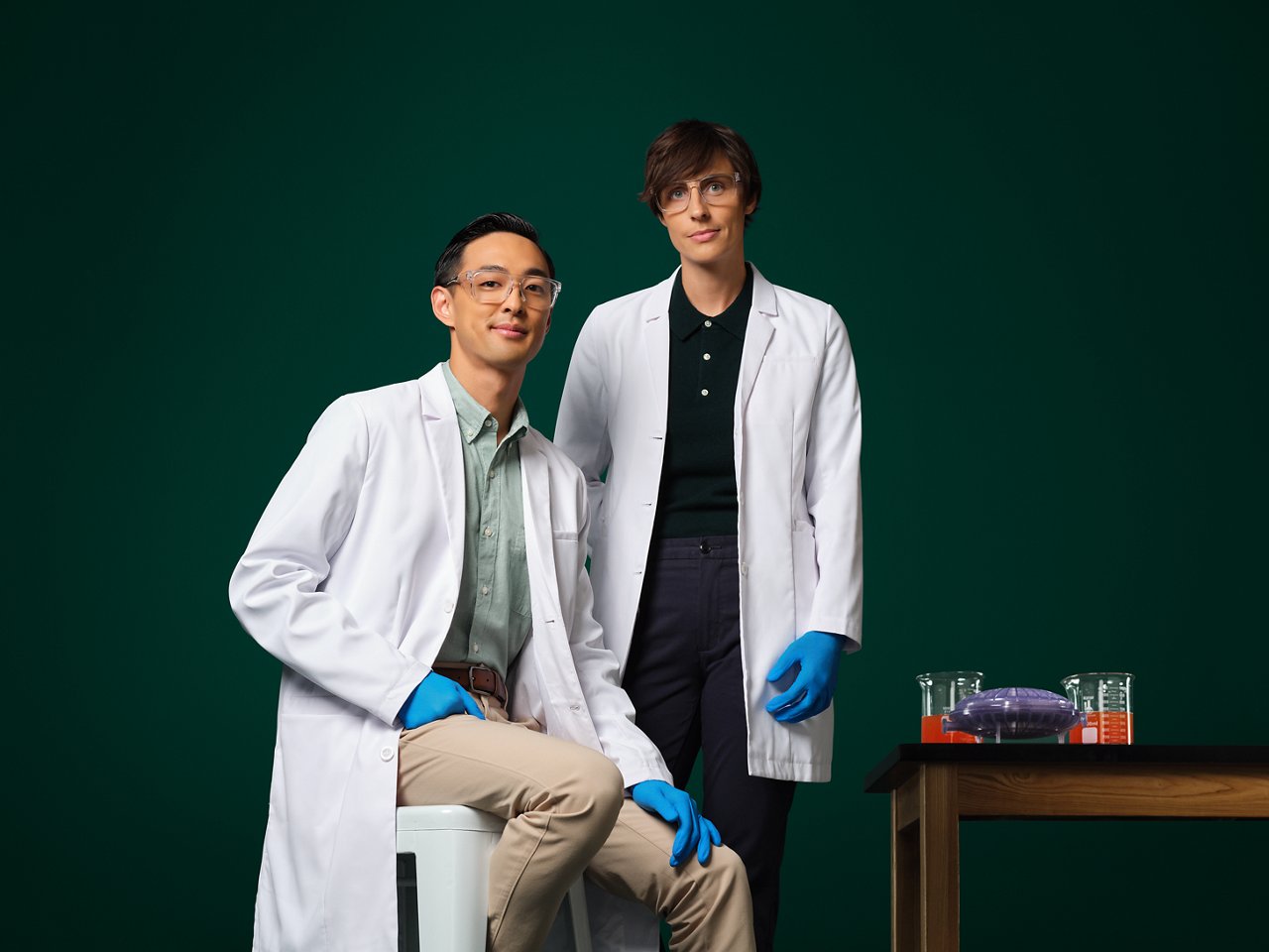 Two engineers in white lab coats and blue gloves by a small table with lab equipment on it against a dark green background.