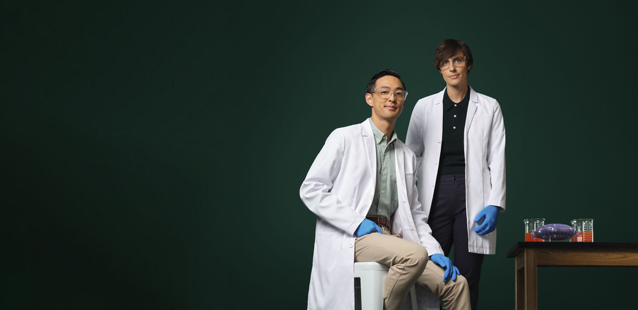 Dos ingenieros con bata blanca y guantes azules junto a una pequeña mesa con material de laboratorio sobre ella