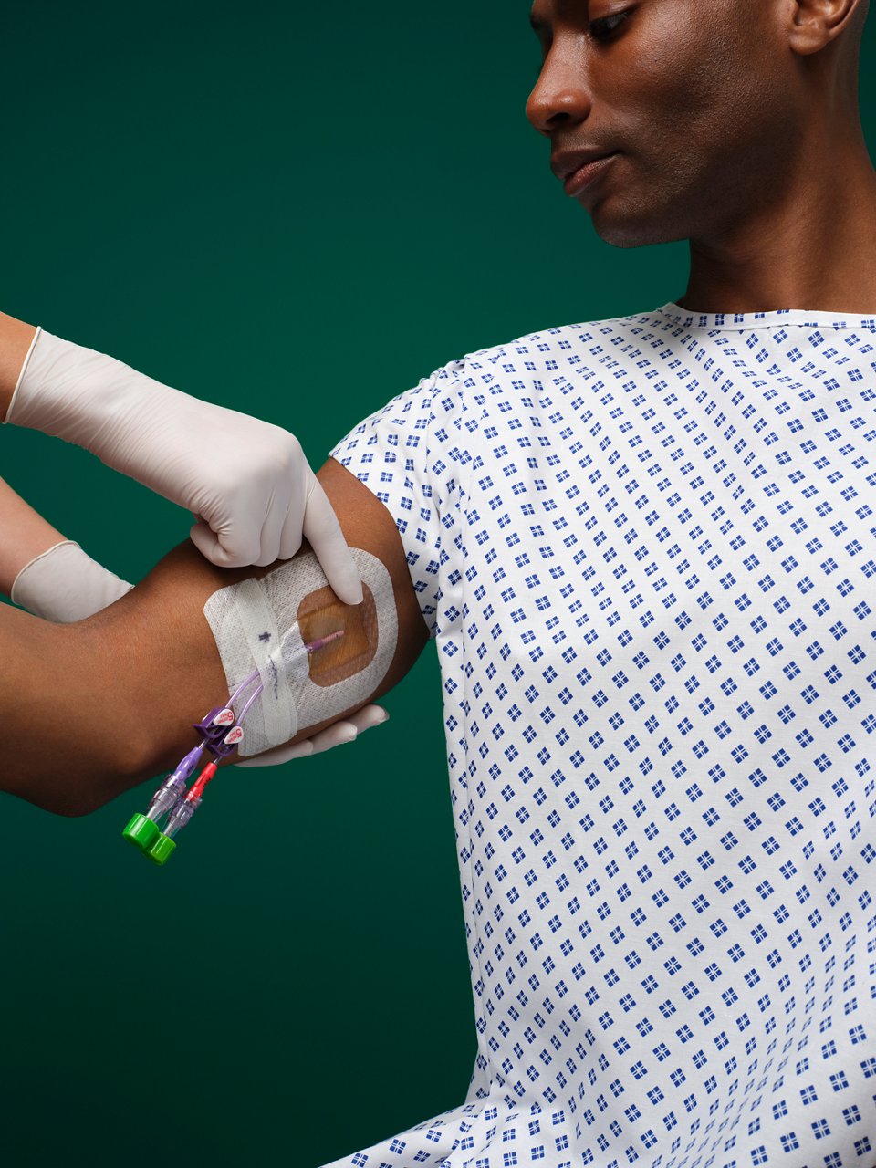 Male patient in hospital gown sits in a dark green room while a 3M Tegaderm CHG I.V. Securement Dressing is applied on his inner bicep.