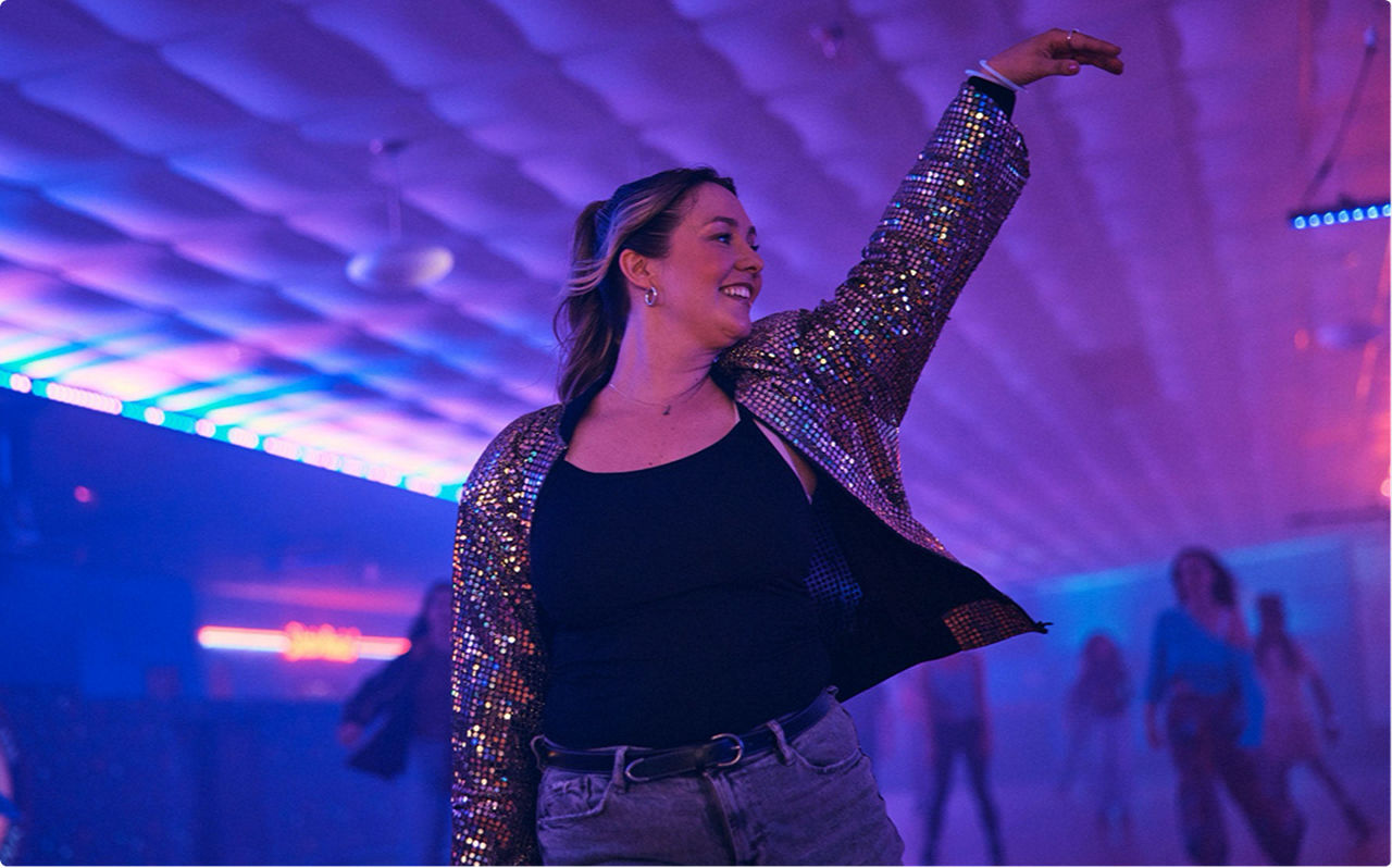 A feminine presenting, light skin toned person roller skating with a smile. Actor portrayals.