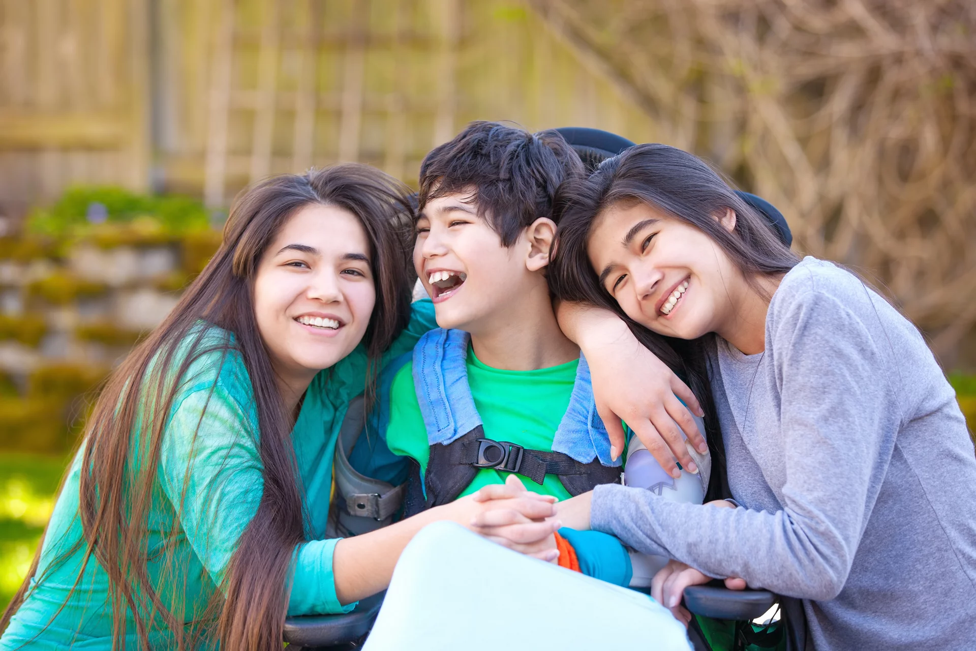 Sisters laughing and hugging disabled little nine year old  brother in wheelchair outdoors