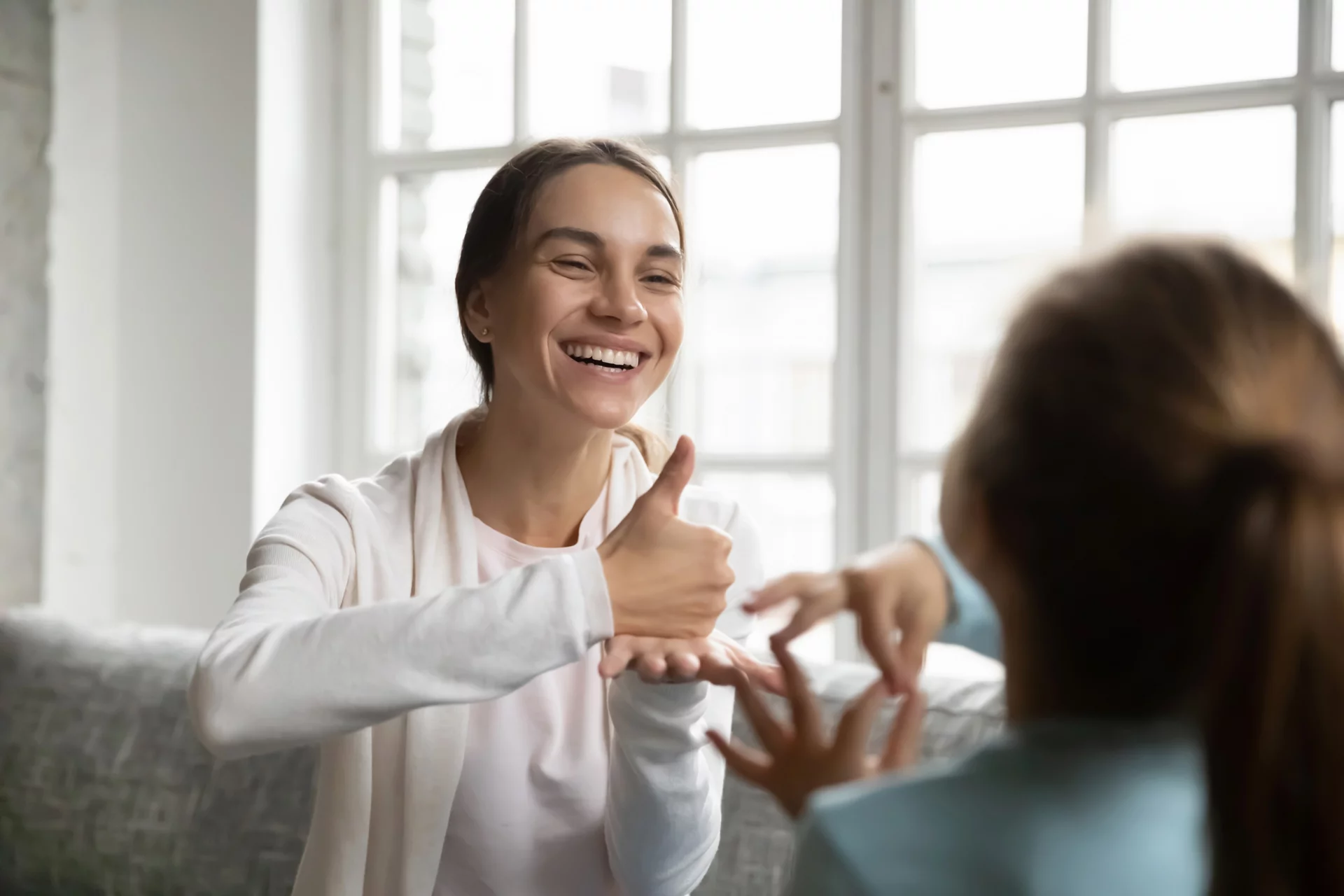 Smiling young Caucasian mom or nanny make hand gesture practice nonverbal talk with little girl child, positive female teacher or tutor learn sign language with small disabled kid, disability concept