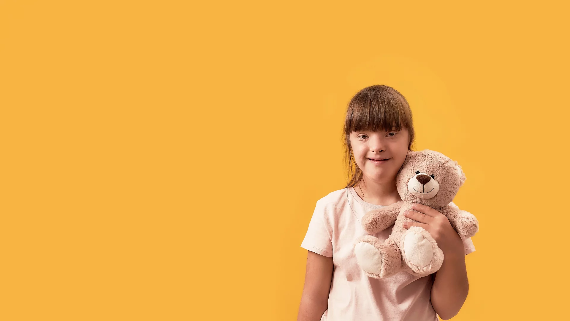 Portrait of disabled girl with Down syndrome smiling at camera, holding her teddy bear while posing isolated over yellow background. Front view. Web Banner