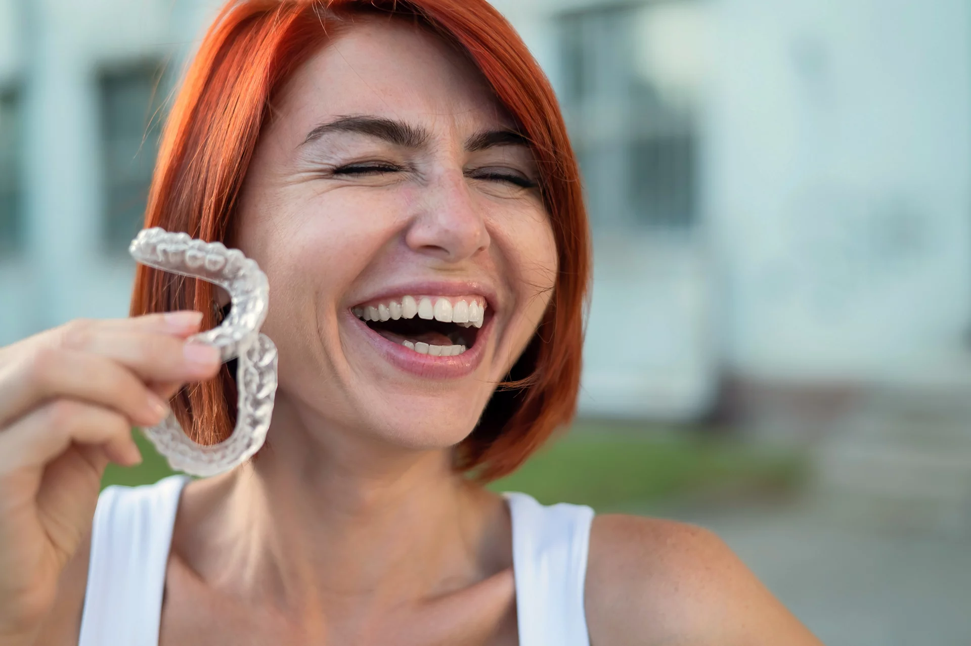 Red-haired Caucasian woman holding transparent mouthguards for bite correction outdoors. A girl with a beautiful snow-white smile uses silicone braces