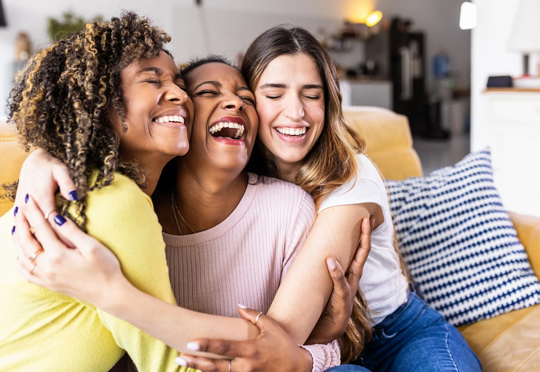 Three happy multiethnic female best friends laughing together - Diverse young women hugging each other and having fun at home - Unity concept
