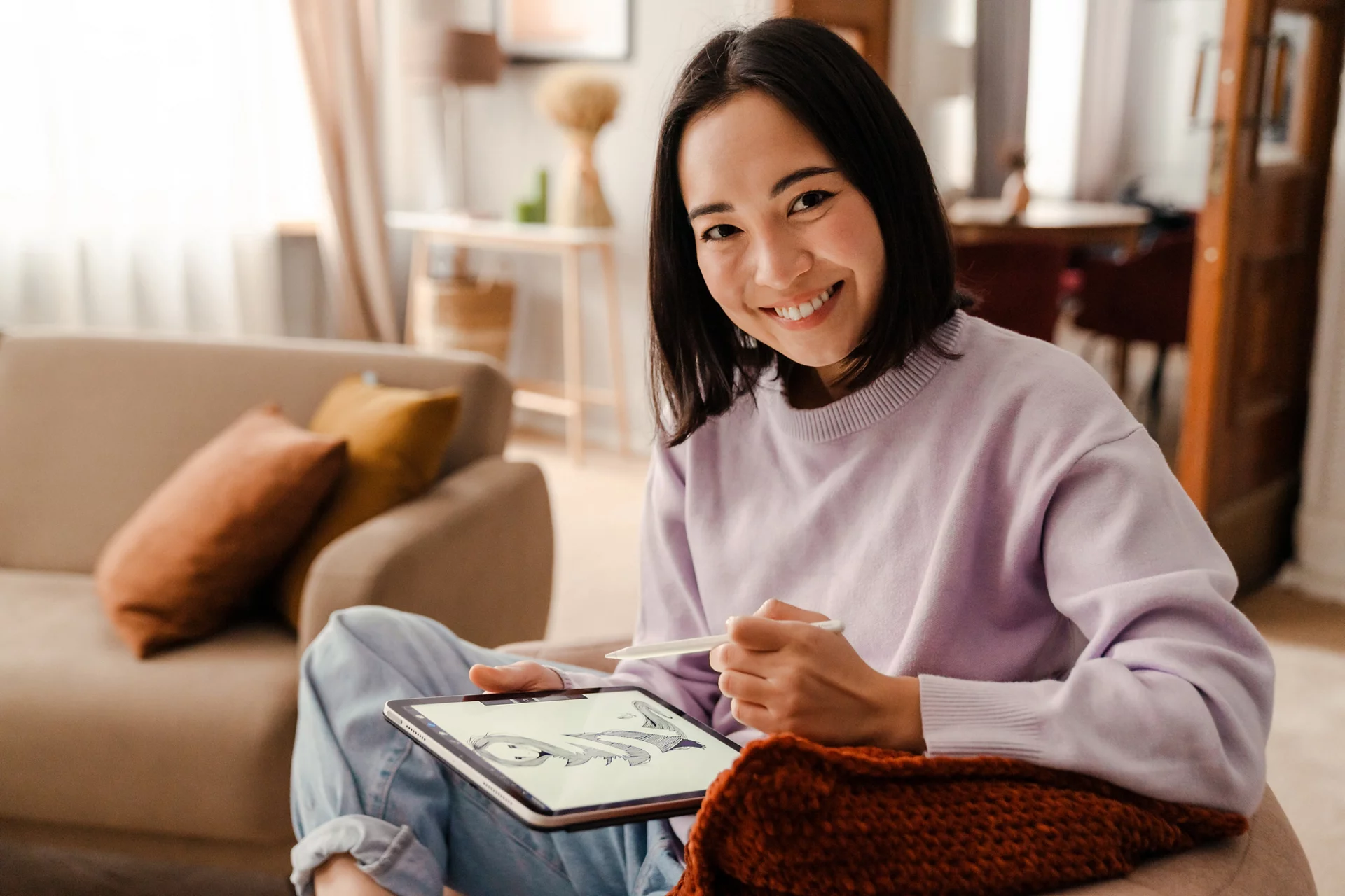 Young beautiful smiling asian woman in casual sweater drawing on digital tablet with stylus while sitting on armchair in cozy living room