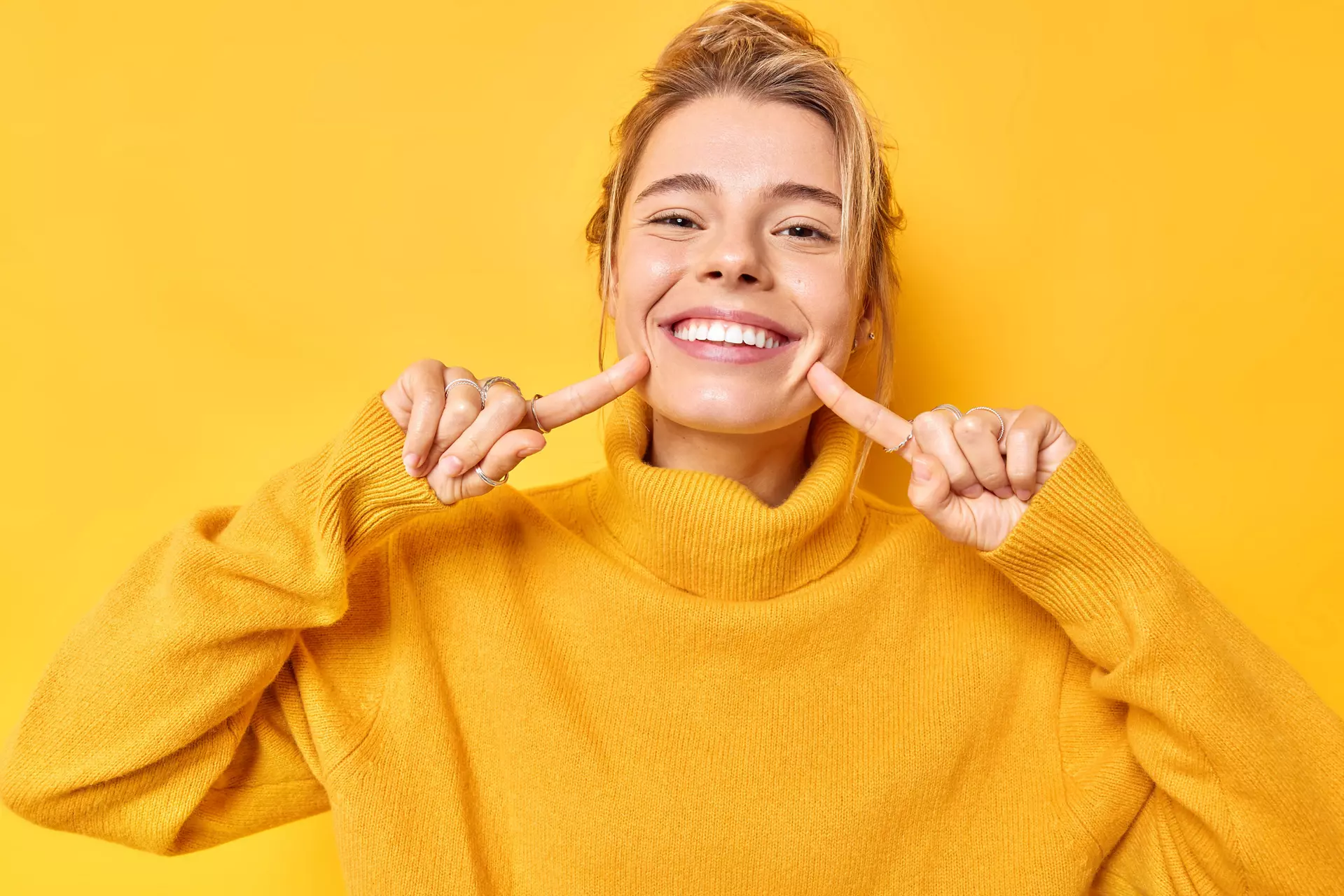 Portrait of cheerful young woman smiles broadly indicates index fingers at face wears casual jumper expresses positive emotions isolated over vivid yellow background. Look at my brilliant smile