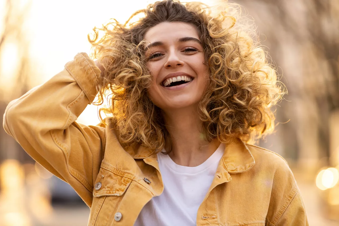 Woman with curley hair and yellow jacket smiling in front of sun