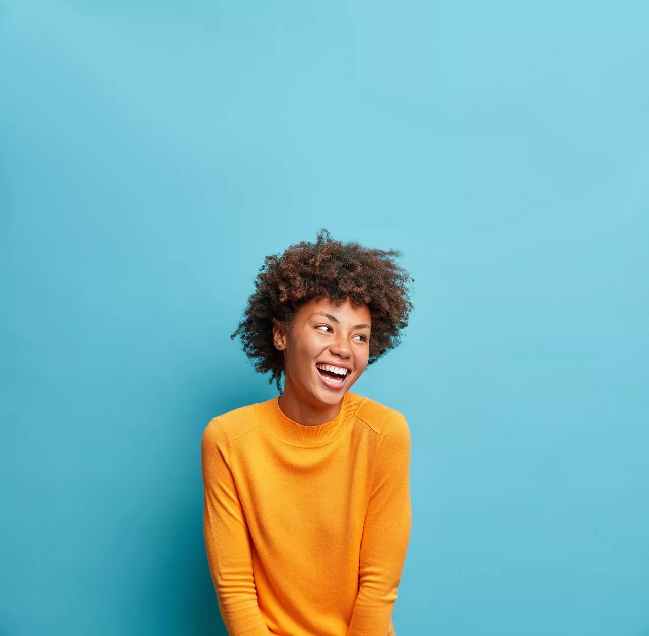 Happy emotions concept. Positive dark skinned beautiful young woman laughs poisitively looks aside with carefree face expression wears casual orange sweater isolated over blue studio background.