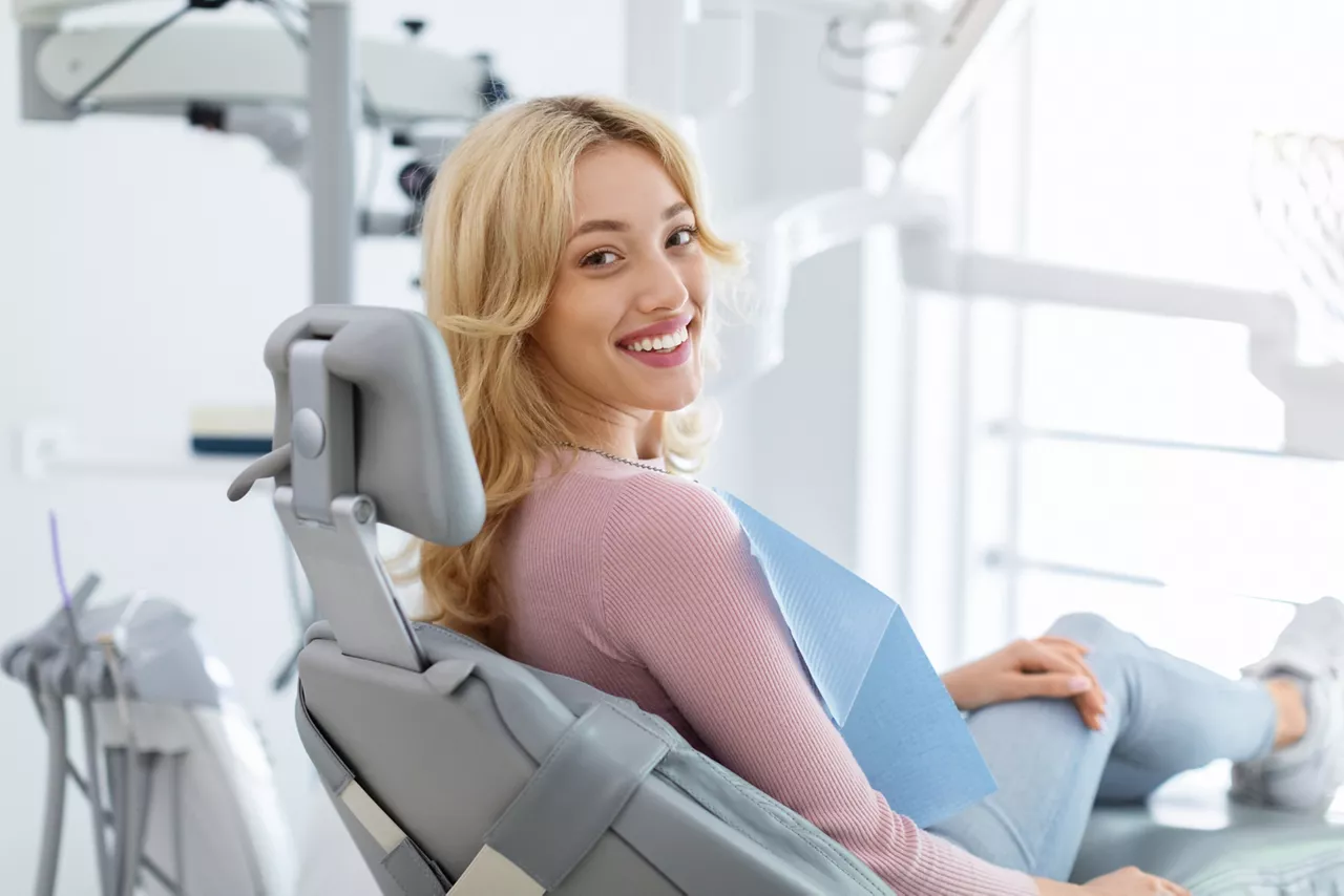 Smiling and relaxed young woman sitting at dental chair in modern clinic, waiting for her dentist. Happy attractive blonde lady attending luxury dental clinic, bacl view, copy space
