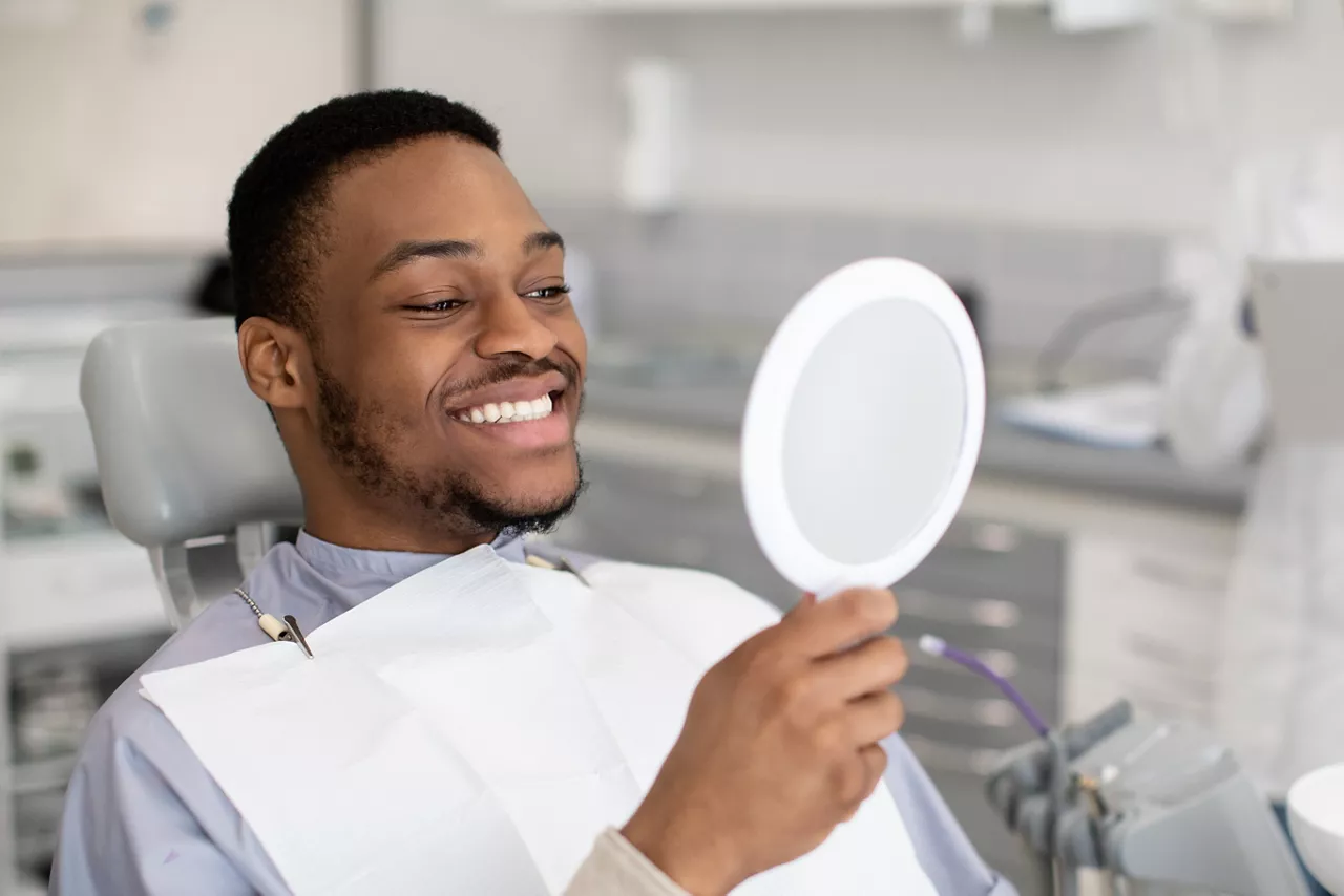 Portrait Of Happy Dental Clinic Patient Looking At Mirror After Treatment, Cheerful African American Guy Sitting In Chair In Stomatological Cabinet And Enjoying His Perfect Smile, Closeup