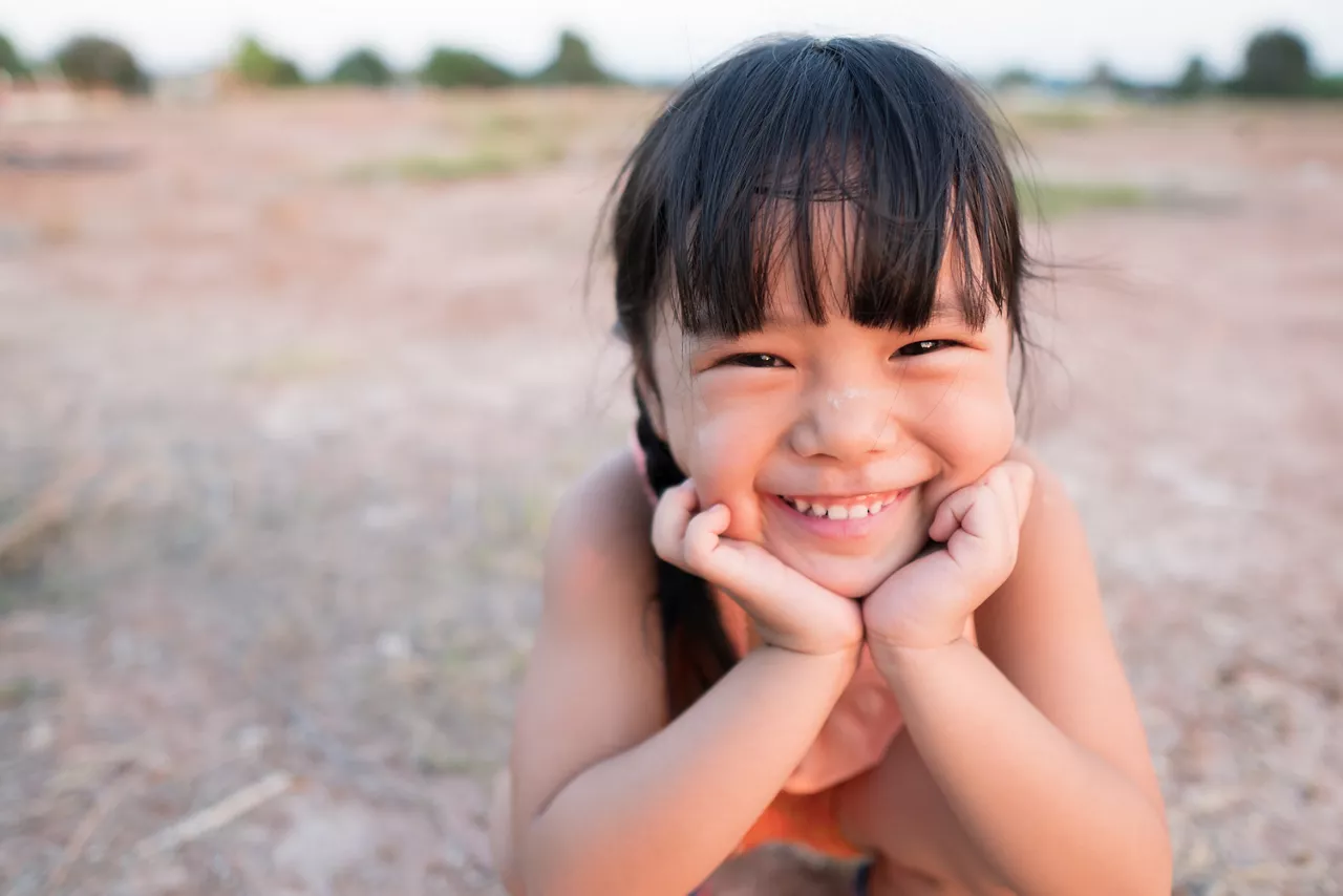 Asian child poor happiness or kid girl smile with laugh and happy fun because come back home to countryside and wear traditional top or sleeveless shirt sit on arid soil field for agriculture at home