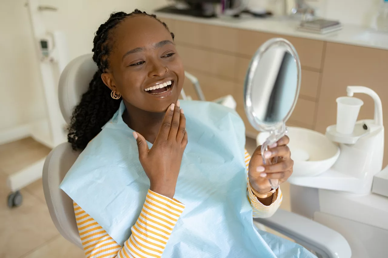 Happy African American female patient looking at her whitened teeth in hand mirror at the dentists'.