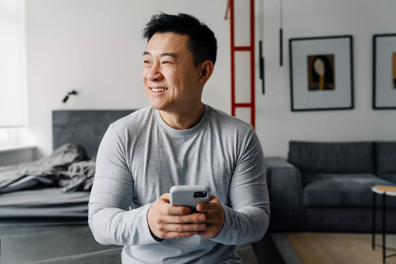 Brunette adult asian man smiling and using cellphone at home