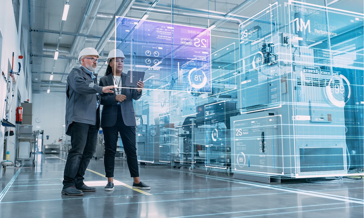 Two factory workers observe data on a screen together, discussing their findings.