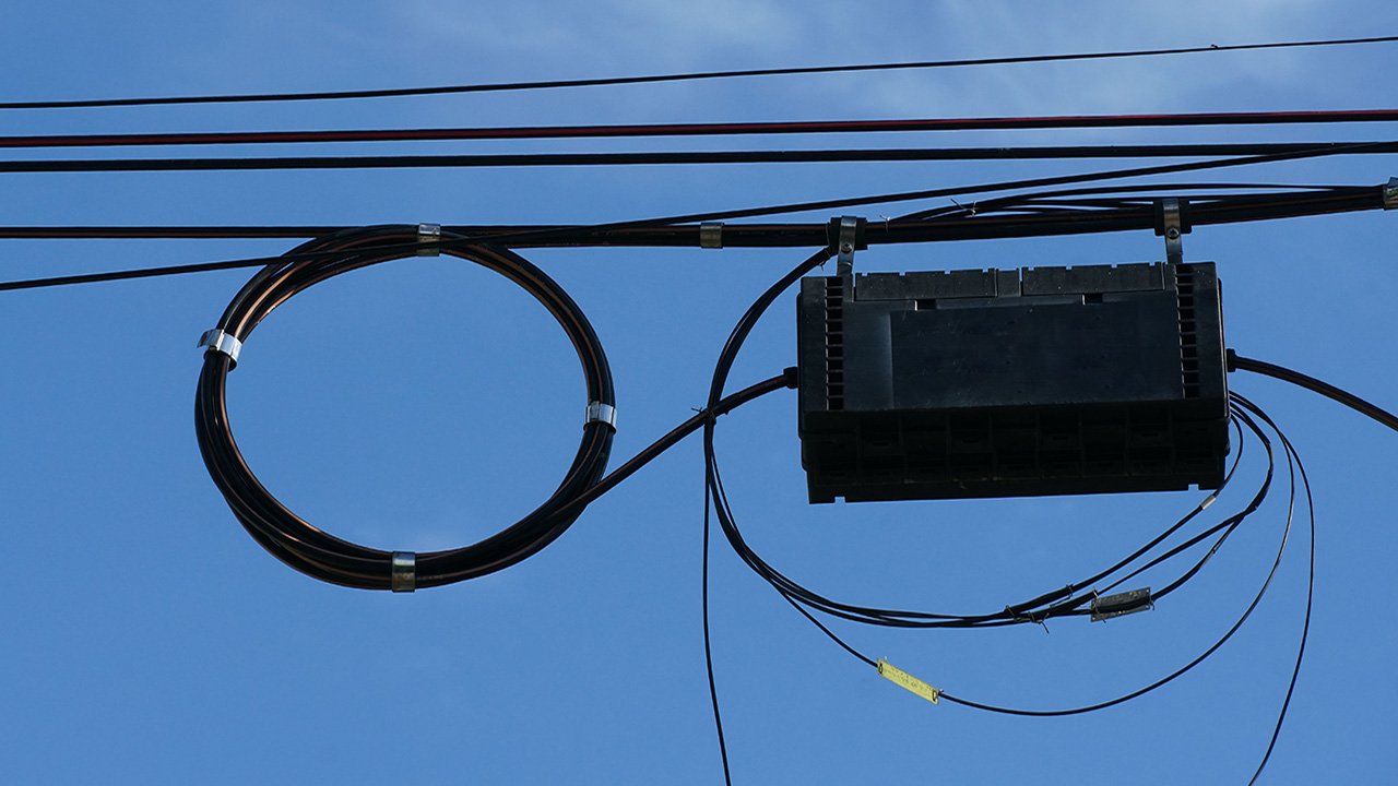 Coil of black fiber cable and a fiber splice box hanging from utility lines against a blue sky in the background 