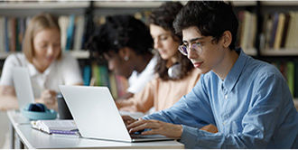 Male student with a blue shirt types on a laptop with an open book alongside and blurred students in the background